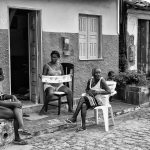 Três mulheres negras estão sentadas em uma cadeira de plástico na rua, mais precisamente em frente à porta de uma casa.